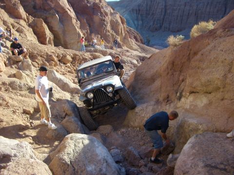 1985 Ford Bronco - A Jeep CJ-7 tackles a challenging off-road course in a desert canyon, with spectators observing the difficult maneuver.