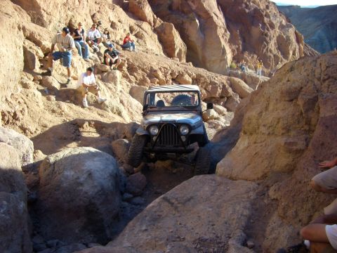 1985 Ford Bronco - A Jeep CJ-7 is driven through a challenging rocky terrain, with spectators observing the off-road adventure.