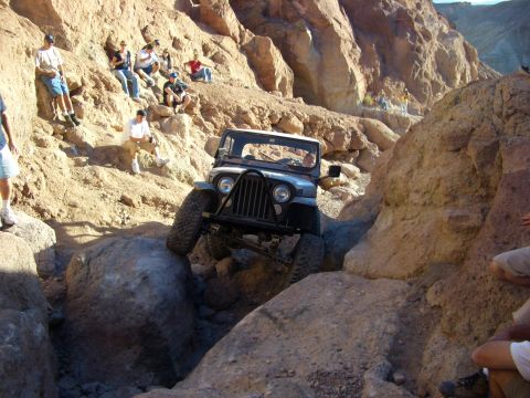 1985 Ford Bronco - A Jeep navigates a difficult rocky trail in a desert canyon, with spectators observing from the slopes.