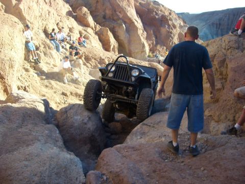 1985 Ford Bronco - A black Jeep Wrangler tackles a challenging off-road course with spectators watching from the surrounding rocks.