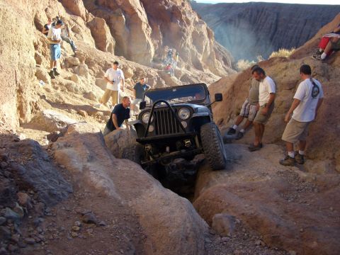 1985 Ford Bronco - A group of people watch as a Jeep Wrangler maneuvers through a narrow, rocky passage in a desert canyon.