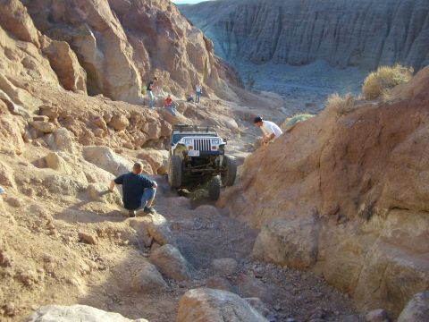 1985 Ford Bronco - A white Jeep Wrangler tackles a challenging off-road trail through a desert canyon, with onlookers observing the ascent.