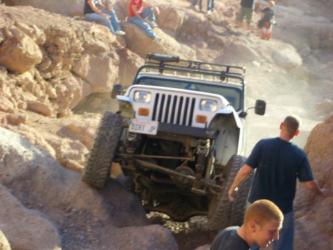 1985 Ford Bronco - A white Jeep Wrangler is shown off-roading on a rocky trail with people watching.