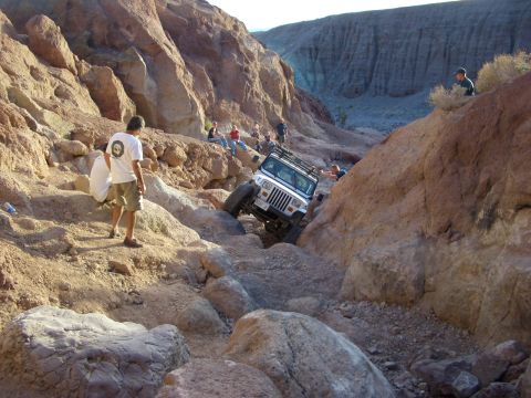 1985 Ford Bronco - A white Jeep Wrangler navigates a challenging off-road trail through a rocky canyon, with spectators observing the ascent.
