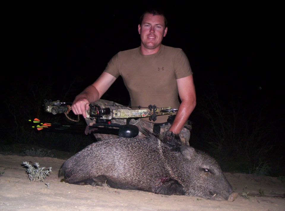 Man in t-shirt and camo pants kneels with a crossbow over a javelina he hunted