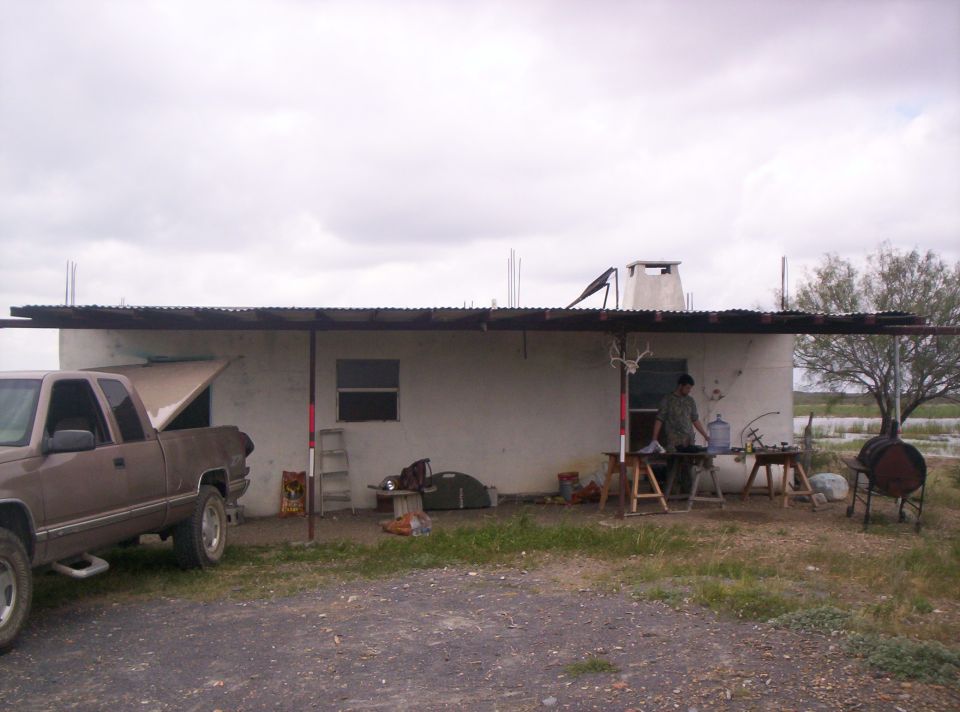 Pickup truck with hood up parked next to a house with an outdoor cooking setup. A person is standing by a grill