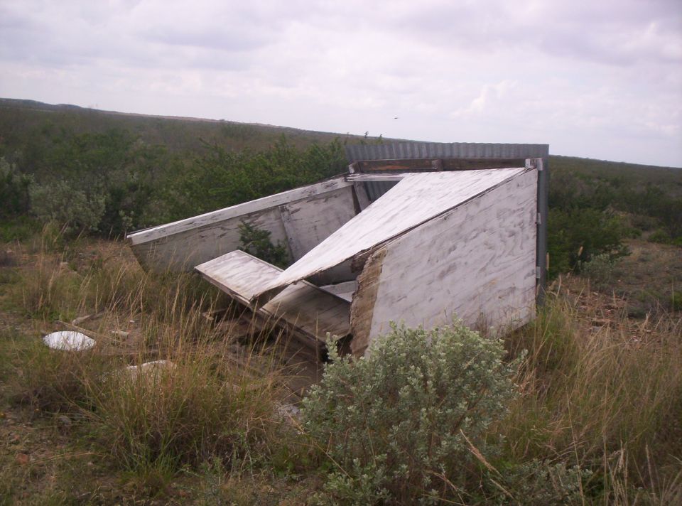 Collapsed wooden and metal structure in a grassy field under a cloudy sky
