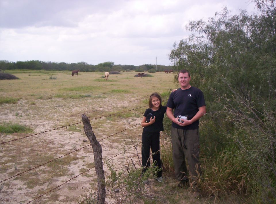 Man and child stand near a barbed wire fence in a grassy field with horses grazing in the distance