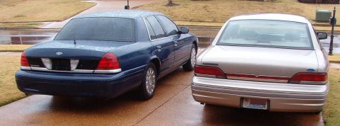 1999 Ford Crown Victoria - Rear view of two Ford Crown Victorias, one blue and one silver, on a wet driveway.