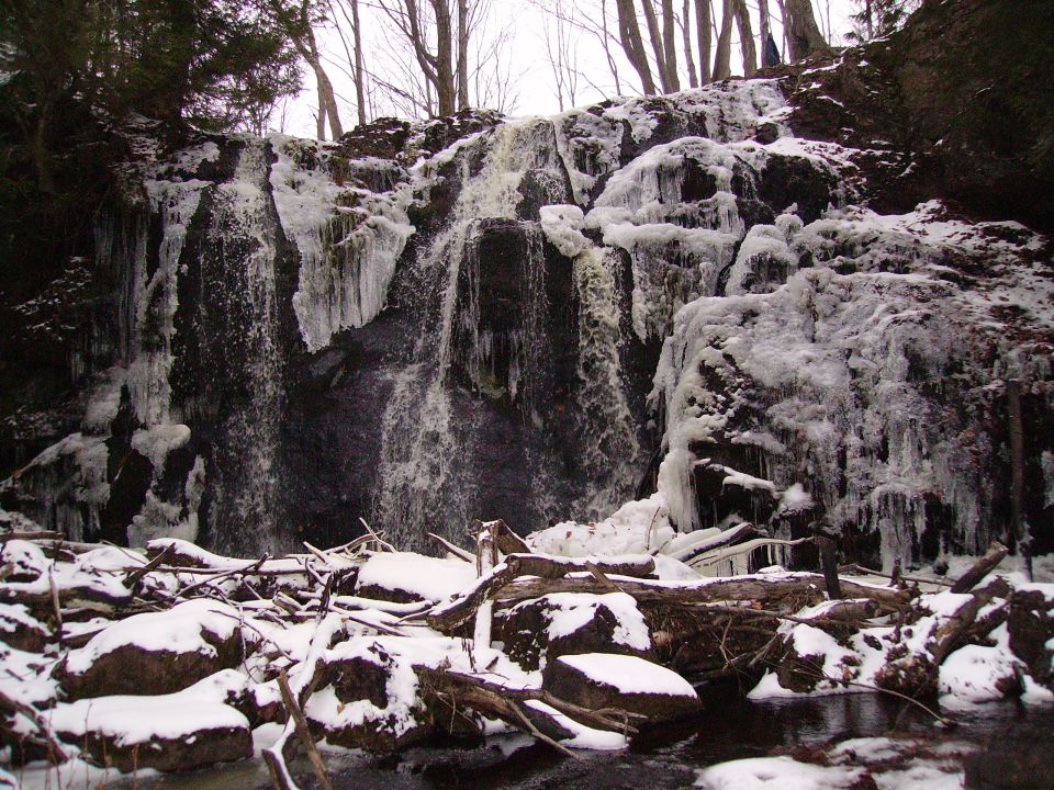 Frozen waterfall with icicles, snow-covered rocks, and fallen logs in the foreground