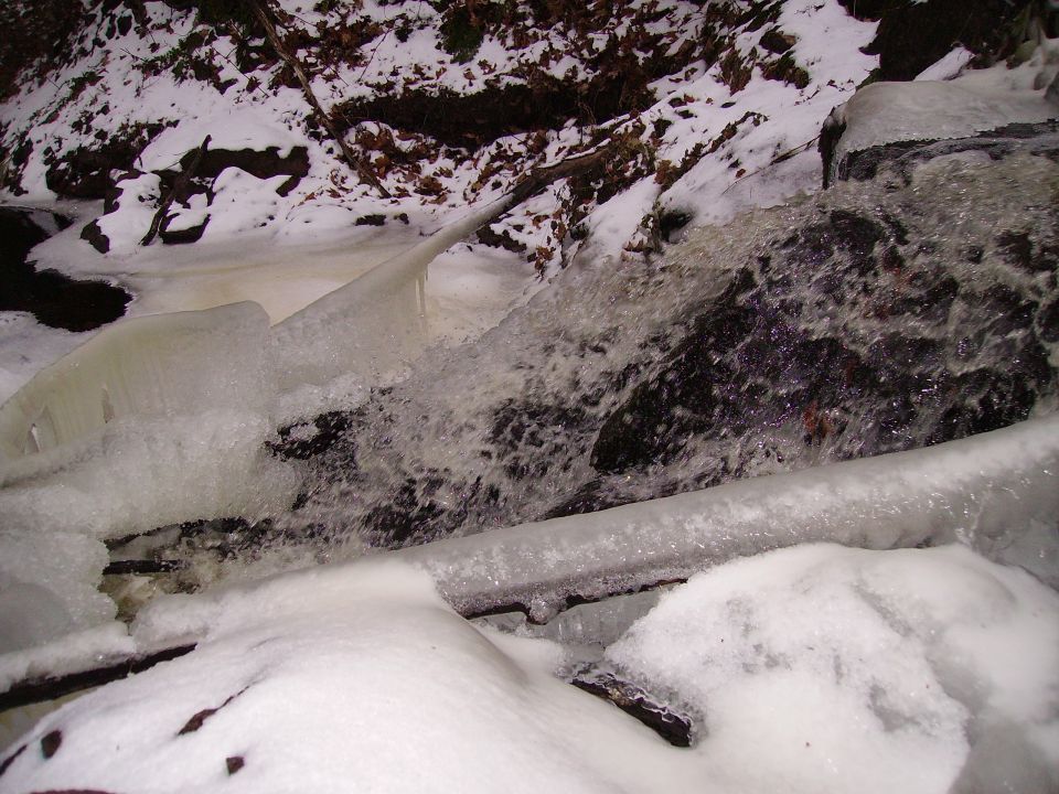 Frozen waterfall with ice formations and rushing water, surrounded by snow-covered rocks and trees