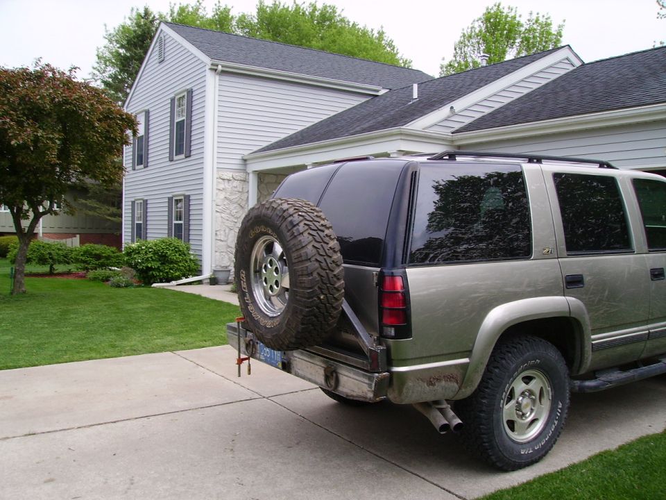 Rear view of a tan SUV with a spare tire mounted on a swing-away carrier. It's parked in a driveway next to a house