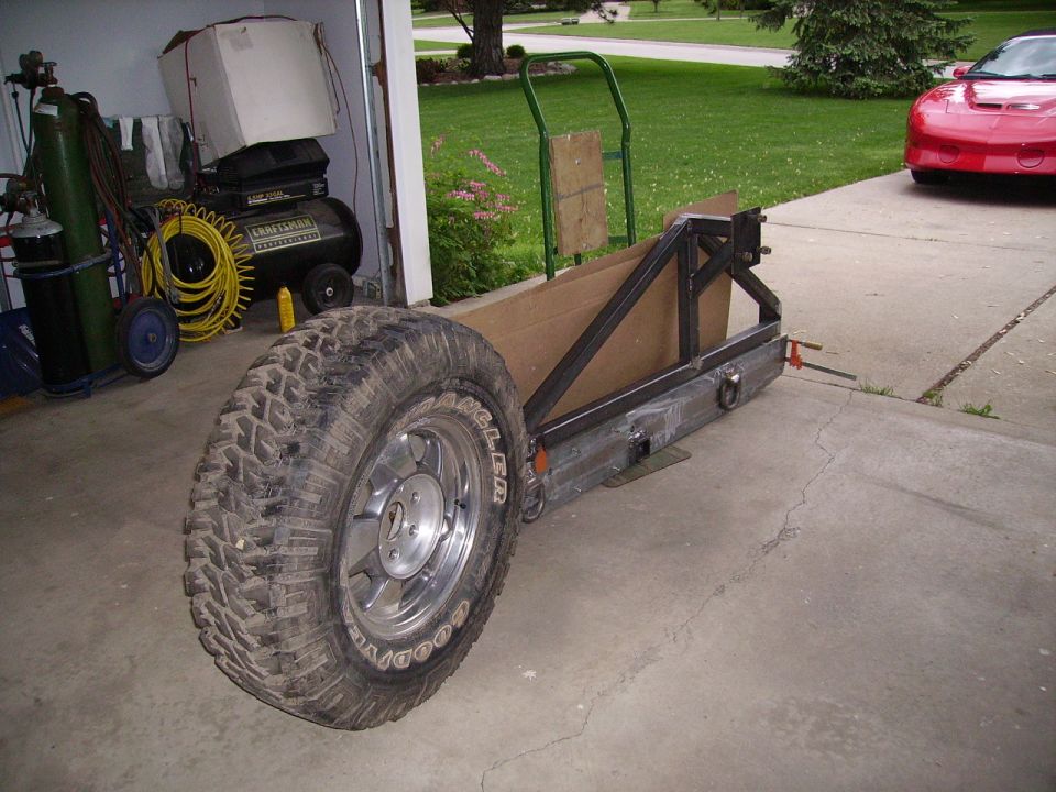 Custom trailer frame with a large off-road tire mounted on the side, parked in a driveway next to a garage
