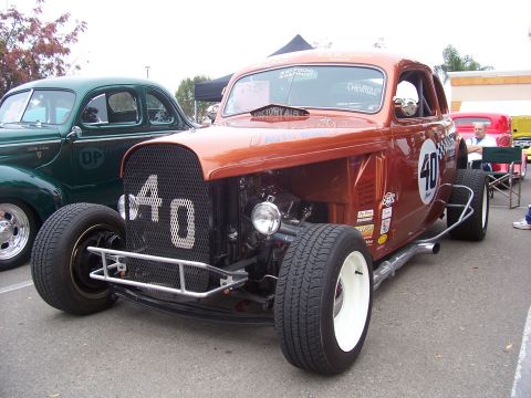 2002 Ford Mustang - A custom hot rod, painted orange, is displayed next to a vintage green car, with a tent and other vehicles in the background.
