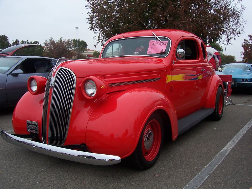Red coupe with chrome bumper and vertical grille. Yellow flames on the side. Another car is parked to the left, and a blue car is in the background