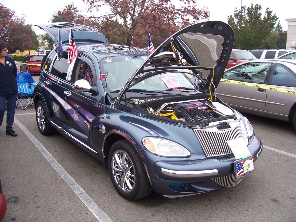 Dark blue PT Cruiser with stars and stripes graphics, hood and trunk open. American flags on poles. Another car is parked nearby