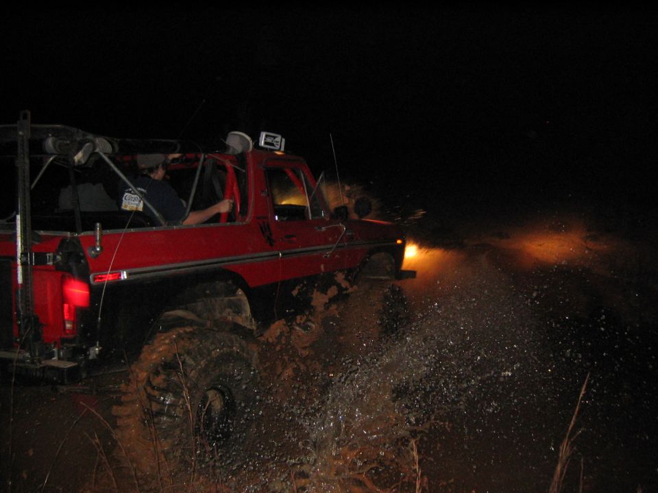 Red 1979 Ford Bronco driving through mud at night, kicking up water
