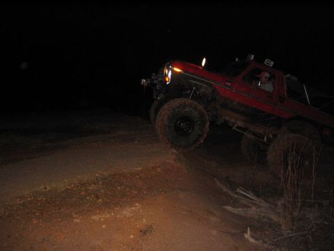 1979 Ford Bronco - A red SUV navigates a dirt path in the dark, its headlights illuminating the terrain.