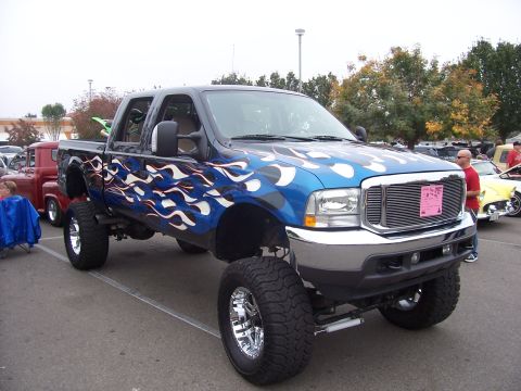 2002 Ford Mustang - A lifted pickup truck with custom flame graphics is displayed at an outdoor event, alongside other vehicles.