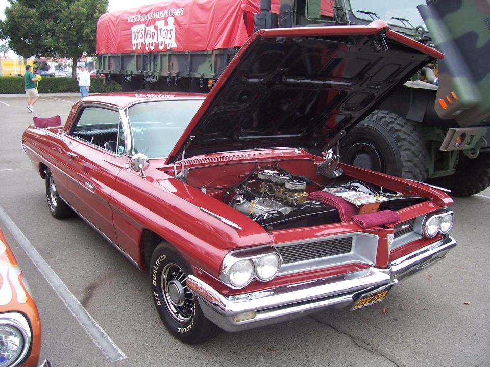 Red 1959 Pontiac Bonneville with hood open, parked next to a military truck