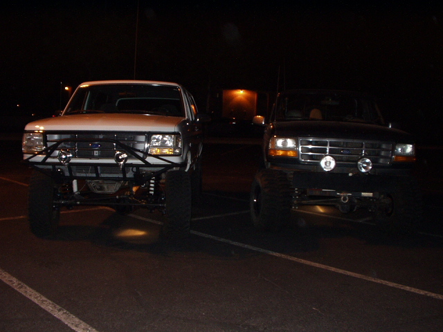 Two lifted Ford trucks parked front-facing at night in a lot