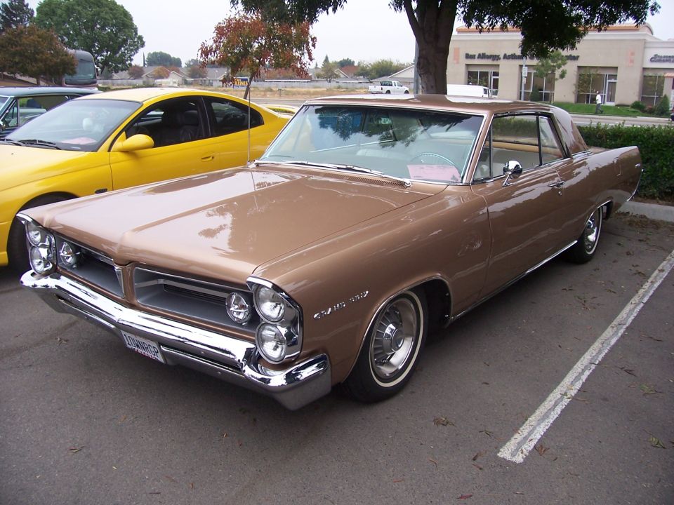 Brown 1963 Pontiac Grand Prix parked next to a yellow car. The Grand Prix has a chrome grille and bumper