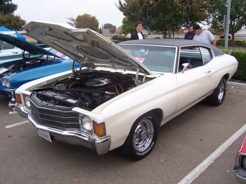 2002 Ford Mustang - A white 1970s Chevrolet Chevelle is displayed with its hood open, parked alongside another vehicle.