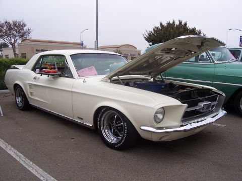 2002 Ford Mustang - A cream 1967 Ford Mustang coupe is parked with its hood open, next to a green vintage car.