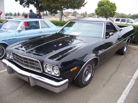 2002 Ford Mustang - A black 1977 Ford Ranchero is parked next to a blue sedan, with trees and houses in the background.