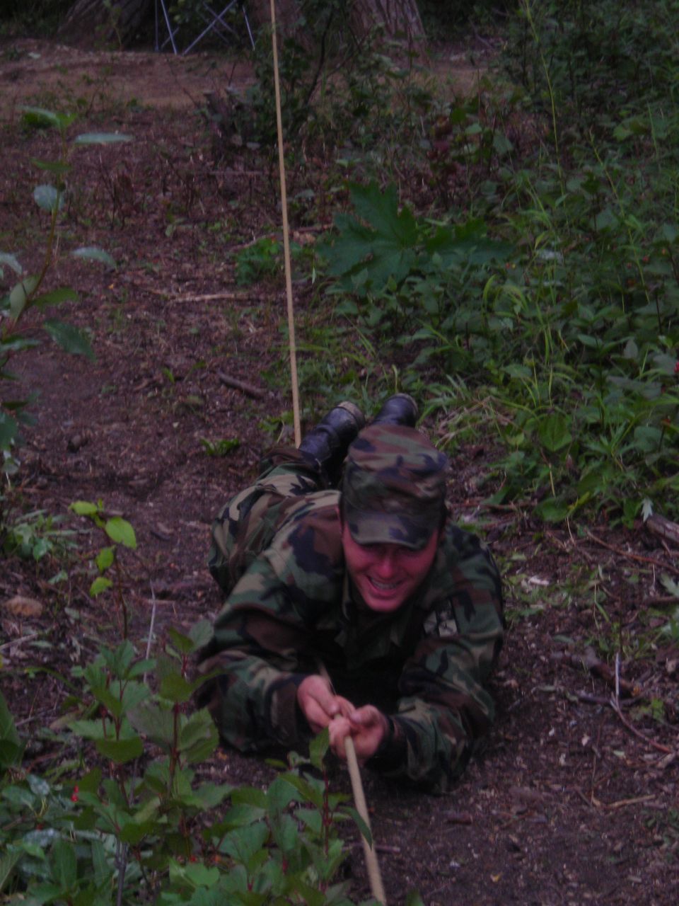 Soldier in camouflage uniform crawling on the ground, pulling a rope. Trees and foliage in background