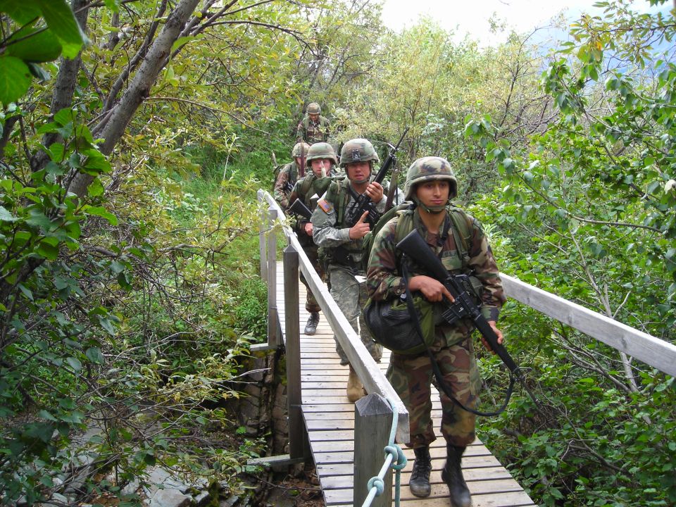 Soldiers in camouflage uniforms and helmets walk single file across a wooden bridge surrounded by trees