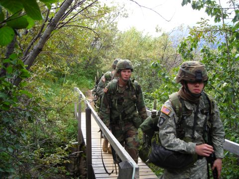 2003 Jeep TJ - Military personnel traverse a narrow wooden bridge surrounded by dense foliage during an outdoor exercise.