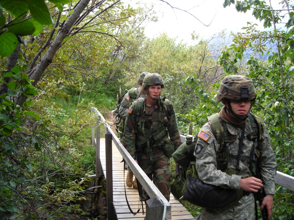 Soldiers in camouflage uniforms walk across a wooden bridge in a wooded area. One soldier has a US flag patch and a 101st Airborne Division patch on his uniform