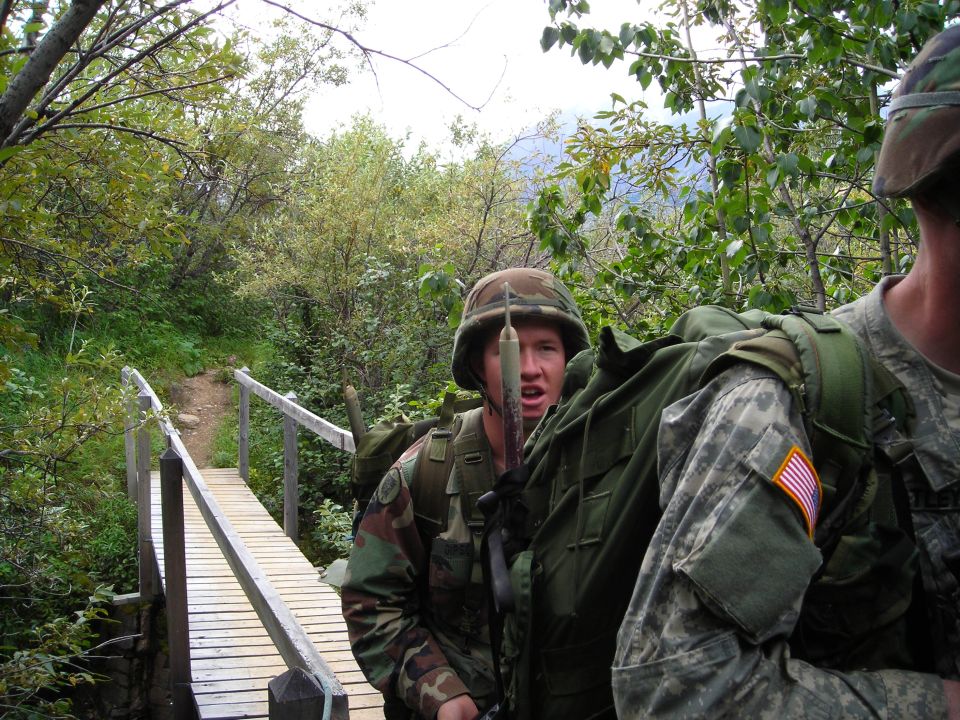 Soldiers in camouflage gear crossing a wooden bridge in a wooded area