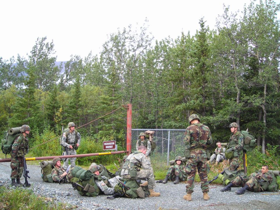 soldiers in camouflage gear with backpacks gathered near a gate and fence, some sitting, some standing