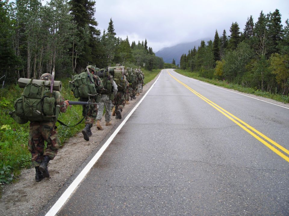 Soldiers marching single file along a paved road with double yellow lines, carrying large backpacks