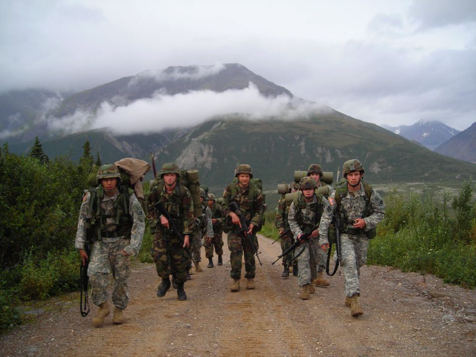 Group of soldiers in camouflage uniforms and helmets marching on a dirt road, carrying backpacks and rifles. Mountains and clouds are visible in the background
