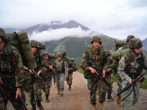 2003 Jeep TJ - A group of soldiers wearing camouflage uniforms and carrying large backpacks march along a dirt path with a mountain range in the distance.
