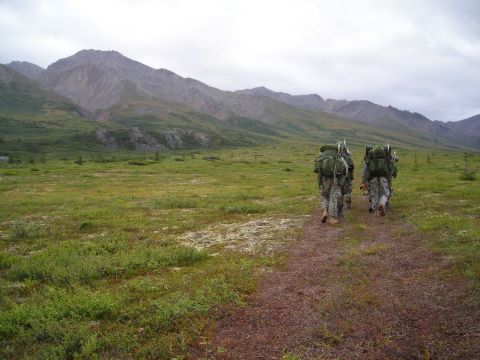 2003 Jeep TJ - Three soldiers with large backpacks walk along a dirt path in a vast, open landscape with mountains in the background.