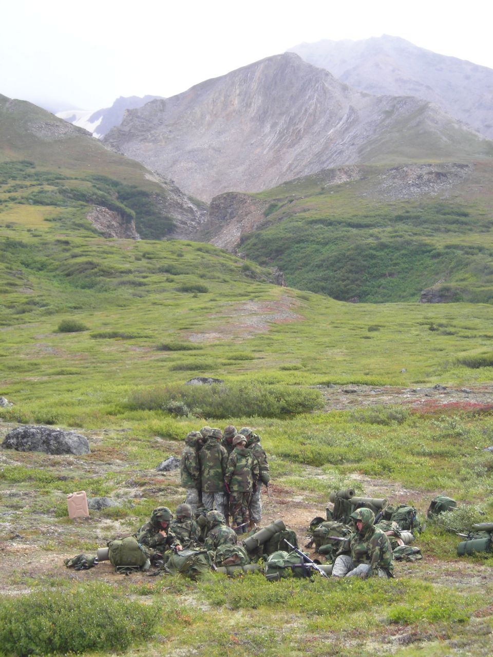 Group of soldiers in camouflage gear gathered in a grassy, mountainous area. Some are standing in a huddle, others are sitting with backpacks