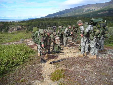 2003 Jeep TJ - A group of soldiers equipped with large backpacks are gathered in a scenic outdoor environment, with trees and hills in the background.