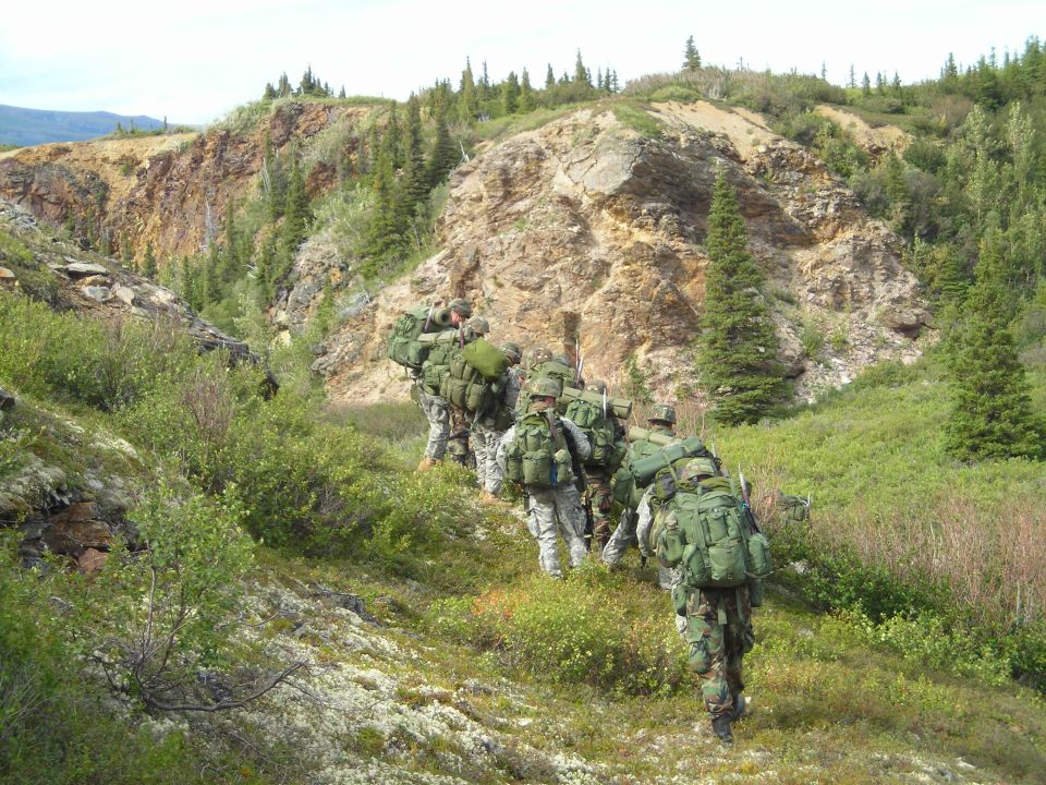 Soldiers in camouflage carrying large backpacks walk up a grassy, rocky incline with sparse trees
