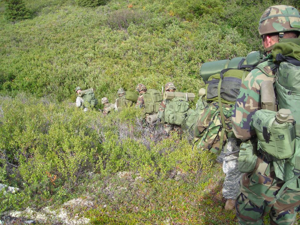 Soldiers in camouflage gear with large backpacks hiking through a green, brushy landscape