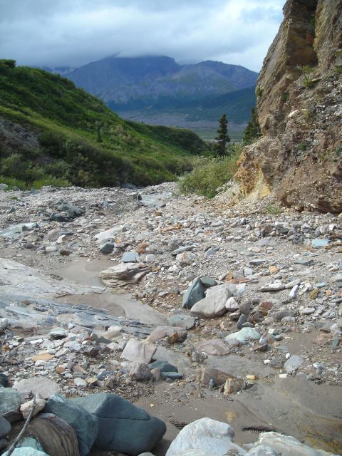 2003 Jeep TJ - A dry, rocky streambed winds through a lush green mountain valley under a cloudy sky.