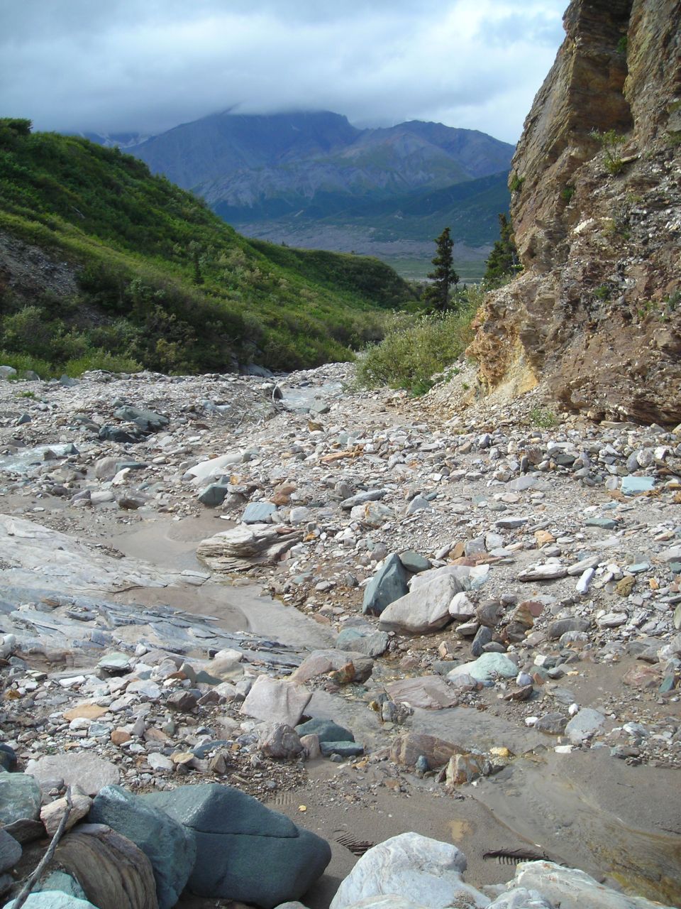 Rocky stream bed in a mountain valley with green hills and a cloudy sky