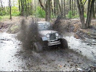 1990 Ford Bronco - A Jeep CJ-5 navigates a challenging muddy path in a wooded area.