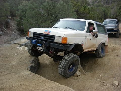 1987 Ford Bronco - A white Ford Bronco navigates a rocky obstacle on an off-road trail, with a second Bronco following.