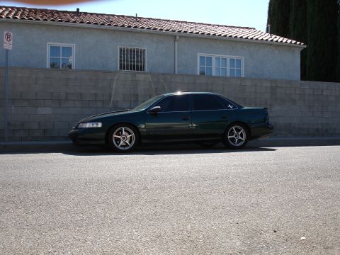 1993 Ford Taurus - Side profile of a dark green sedan with aftermarket wheels parked on a street.