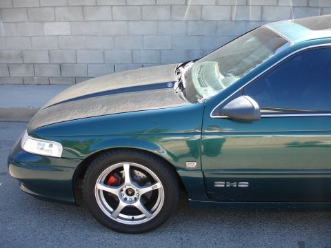 1993 Ford Taurus - A green sedan is parked in front of a brick wall, showing its front wheel and side profile.