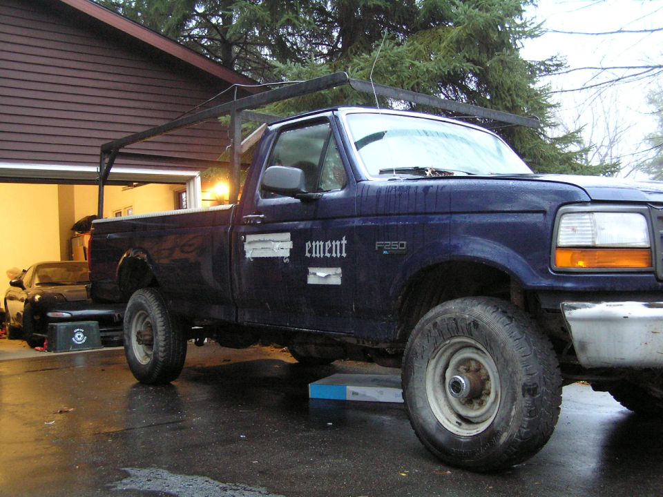 Dark blue Ford F-250 truck with a rack, parked in front of a garage. A black sports car is visible inside the garage